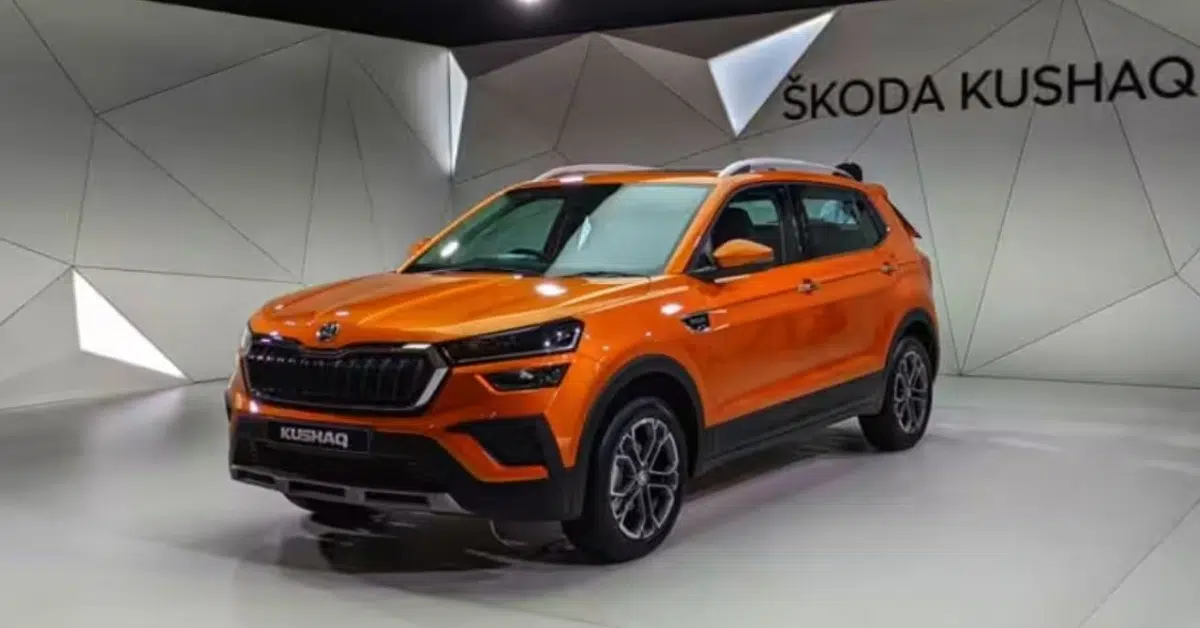 Orange Škoda Kushaq SUV displayed indoors, shown from a front three-quarter angle with black grille, LED headlights, alloy wheels, and roof rails against a geometric studio backdrop.